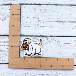 May include: Embroidered patch featuring a dog dressed as a ghost, holding a jack-o'-lantern. The patch is white, tan, and orange, with black outlining. The patch is approximately 1 inch in height and 1.5 inches in length. The background is a white wooden surface.