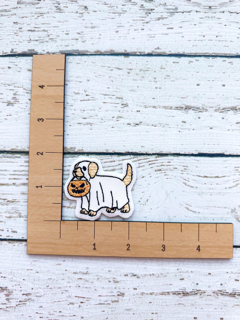 May include: Embroidered patch featuring a dog dressed as a ghost, holding a jack-o'-lantern. The patch is white, tan, and orange, with black outlining. The patch is approximately 1 inch in height and 1.5 inches in length. The background is a white wooden surface.