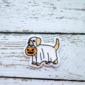 May include: Embroidered patch featuring a white dog wearing a ghost costume and carrying a jack-o'-lantern. The patch has a white background with black and tan accents, perfect for Halloween.