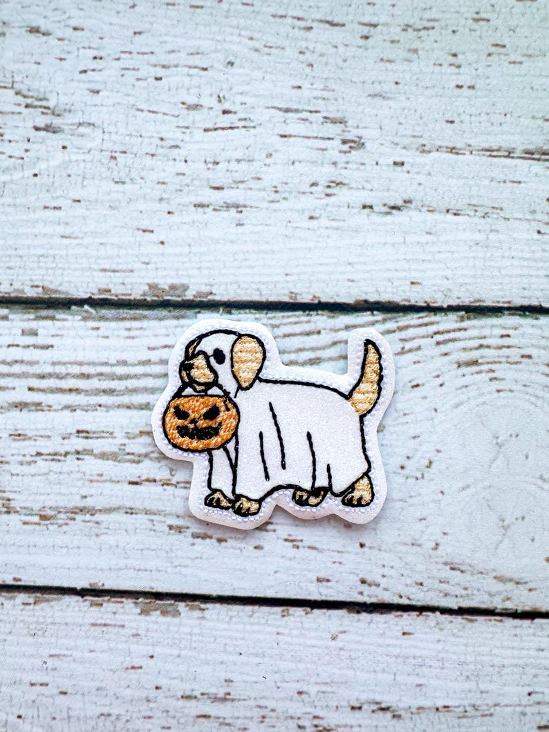 May include: Embroidered patch featuring a white dog wearing a ghost costume and carrying a jack-o'-lantern. The patch has a white background with black and tan accents, perfect for Halloween.