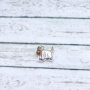 May include: A Halloween-themed charm featuring a white dog wearing a ghost costume and carrying a jack-o'-lantern. The charm has a white border and is set against a white wooden background.