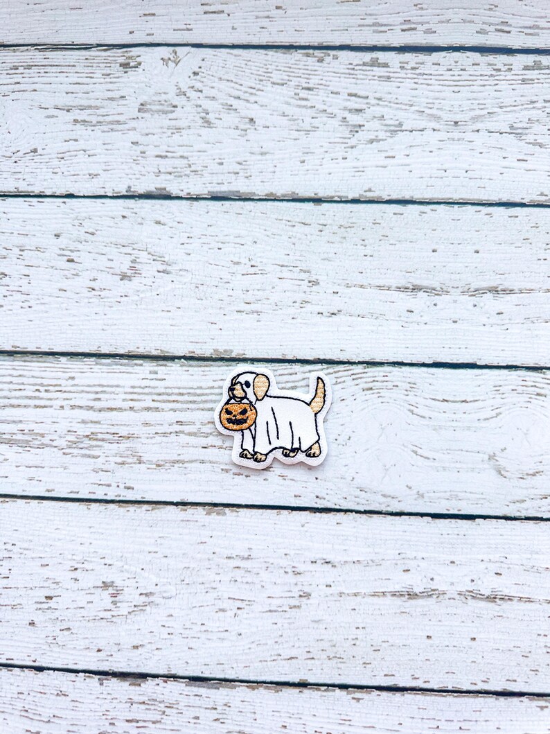 May include: A Halloween-themed charm featuring a white dog wearing a ghost costume and carrying a jack-o'-lantern. The charm has a white border and is set against a white wooden background.