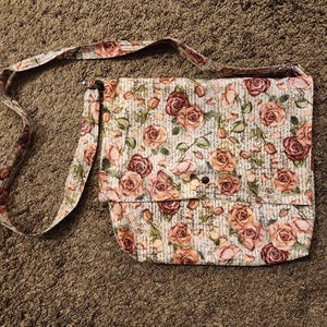 以下が含まれることがあります： ピンクとブラウンのバラの花柄が施された白い布製のショルダーバッグ。バッグには長いストラップとフラップクロージャーが付いています。
