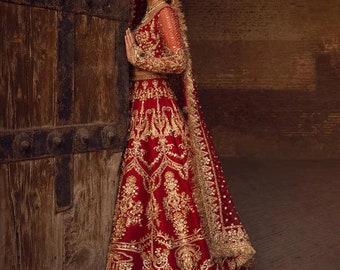 May include: A woman wearing a red and gold embroidered lehenga with a matching dupatta. The lehenga has intricate floral designs and is paired with a red blouse. The woman is standing in front of a wooden door.