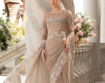 May include: A beige saree with intricate embroidery and embellishments. The saree has a sheer overlay with a delicate lace trim. The model is wearing a matching blouse with a plunging neckline and cold shoulder sleeves.