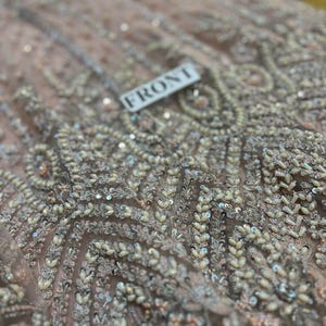 May include: Close-up of a light pink fabric with intricate beading and sequins. The fabric has a pattern of small, white beads arranged in a geometric design. The word "FRONT" is written on a white tag.