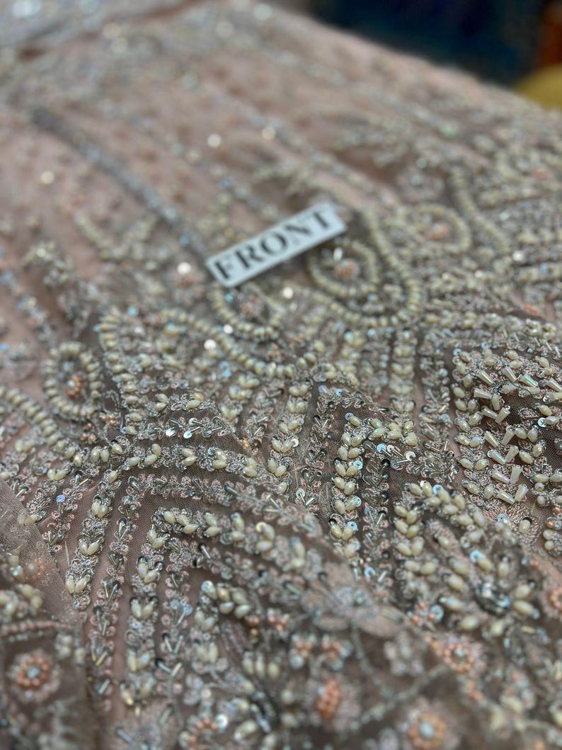 May include: Close-up of a light pink fabric with intricate beading and sequins. The fabric has a pattern of small, white beads arranged in a geometric design. The word "FRONT" is written on a white tag.