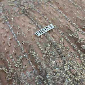 May include: Close-up of a light pink fabric with intricate beading and sequins. The fabric has a delicate, floral pattern and is adorned with small, white beads and silver sequins. The fabric is labeled "FRONT".