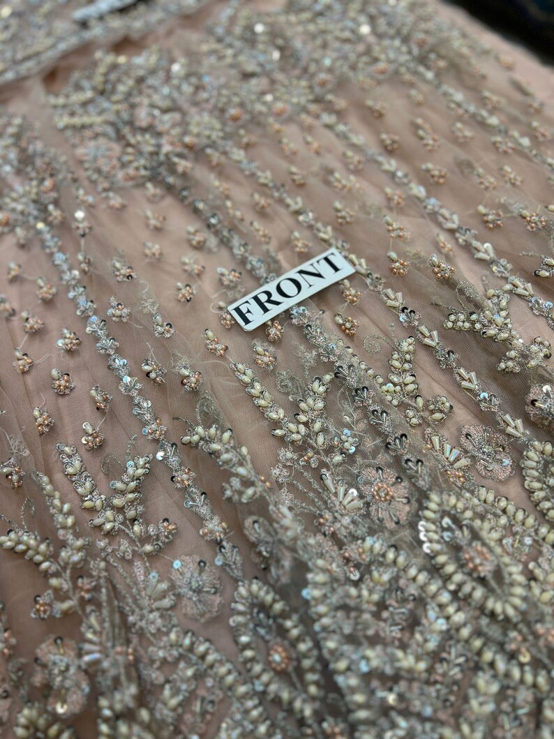 May include: Close-up of a light pink fabric with intricate beading and sequins. The fabric has a delicate, floral pattern and is adorned with small, white beads and silver sequins. The fabric is labeled "FRONT".