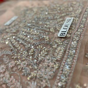 May include: Close-up of a light brown fabric with intricate silver and white beading and sequins. The fabric has a delicate, sheer texture. The word "BLOUSE" is written on a white tag.