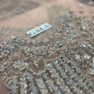 May include: Close-up of a light pink fabric with intricate silver embroidery. The fabric has a delicate, net-like texture and is adorned with sequins and beads. A white label with the word "GALA" is visible in the upper right corner.