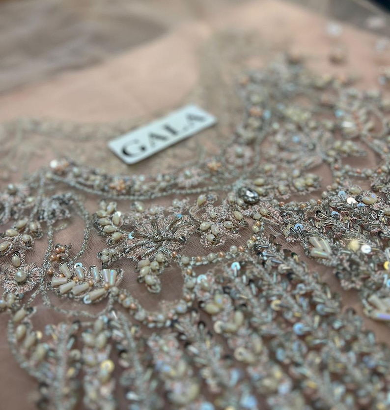 May include: Close-up of a light pink fabric with intricate silver embroidery. The fabric has a delicate, net-like texture and is adorned with sequins and beads. A white label with the word "GALA" is visible in the upper right corner.