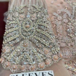 May include: Close-up of a light pink fabric with intricate silver embroidery and beading. The fabric has a delicate, sheer texture and features a floral design. The text "SLEEVES" is visible in the lower right corner of the image.