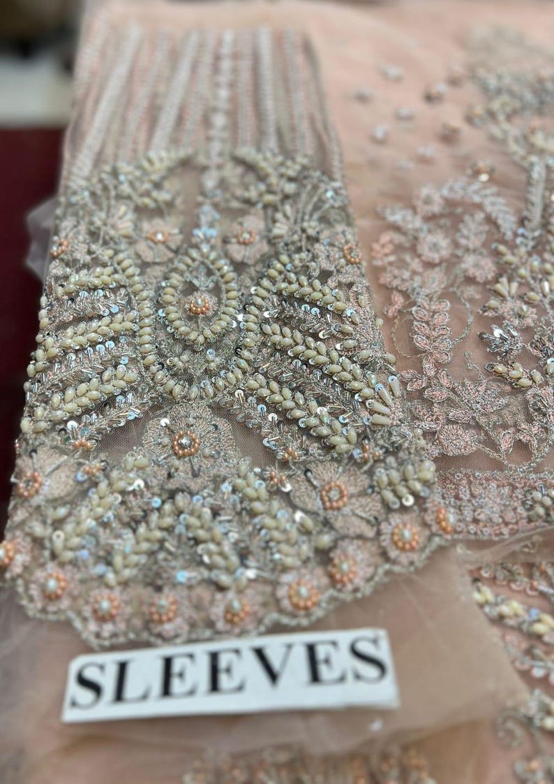 May include: Close-up of a light pink fabric with intricate silver embroidery and beading. The fabric has a delicate, sheer texture and features a floral design. The text "SLEEVES" is visible in the lower right corner of the image.