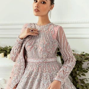 May include: A woman wearing a pink and silver embellished gown with a long, flowing skirt. The gown has a high neckline and long sleeves. The woman is wearing a ring on her finger and earrings.