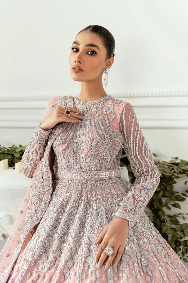 May include: A woman wearing a pink and silver embellished gown with a long, flowing skirt. The gown has a high neckline and long sleeves. The woman is wearing a ring on her finger and earrings.