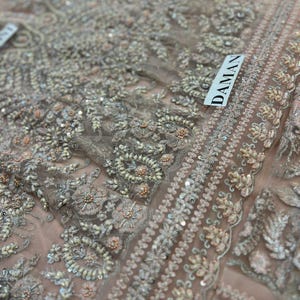 May include: Close-up of a delicate, light pink fabric with intricate embroidery and sequins. The fabric has a floral pattern with small pearls and beads. The word "DAMAN" is visible in the upper right corner.