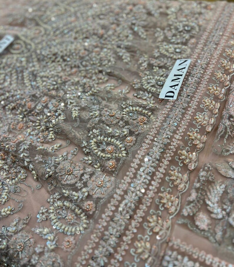 May include: Close-up of a delicate, light pink fabric with intricate embroidery and sequins. The fabric has a floral pattern with small pearls and beads. The word "DAMAN" is visible in the upper right corner.