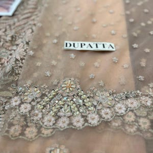 May include: Close-up of a light brown sheer fabric with intricate floral embroidery and pearl embellishments. The fabric has a label that reads "DUPATTA".