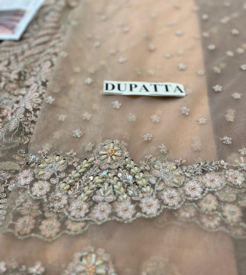 May include: Close-up of a light brown sheer fabric with intricate floral embroidery and pearl embellishments. The fabric has a label that reads "DUPATTA".