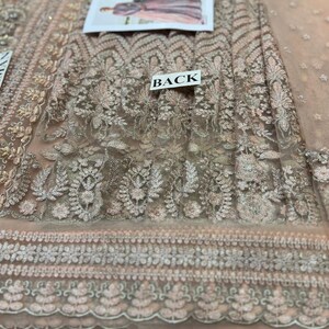 May include: A close-up of a light pink sheer fabric with intricate silver embroidery. The fabric has a floral pattern and a scalloped edge. The word "BACK" is printed on a white tag.