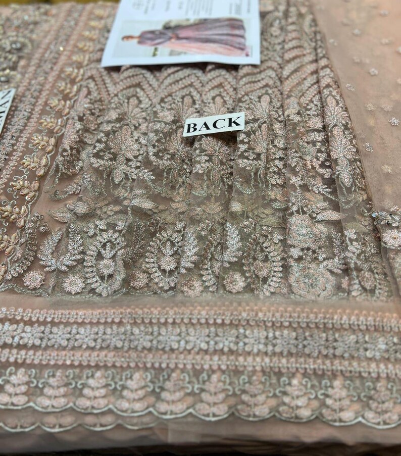 May include: A close-up of a light pink sheer fabric with intricate silver embroidery. The fabric has a floral pattern and a scalloped edge. The word "BACK" is printed on a white tag.