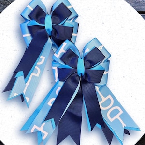 May include: Two blue and navy equestrian-themed bows. The bows feature a light blue ribbon with a white equestrian bit pattern, and a dark blue ribbon. The bows are layered and tied with a light blue ribbon in the center.