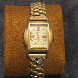 Puede incluir: Un reloj de pulsera dorado con una esfera blanca y una correa dorada. El reloj tiene una esfera rectangular con un pequeño segundero y una ventana de fecha. La esfera del reloj tiene algunos pequeños diamantes alrededor del borde y algunas piedras rojas en la parte superior.