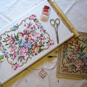 May include: A cross-stitch embroidery project in progress, featuring a floral design with pink, yellow, and blue flowers. The embroidery is mounted in a wooden frame and includes a printed pattern with the word "ASTORIA".