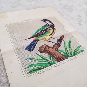 May include: A vintage cross-stitch pattern featuring a green, yellow, and blue bird perched on a brown branch with green leaves. The pattern is printed on a white paper with the text "&agrave; Paris chez Mallez, A&icirc;n&eacute; Editeur, Rue St. George, N&deg;29".