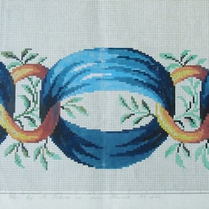 May include: A needlepoint design featuring three linked blue orbs with a textured appearance. The orbs are connected by a ribbon in shades of orange and yellow, with green leafy sprigs interspersed throughout. The background is a light, off-white fabric.