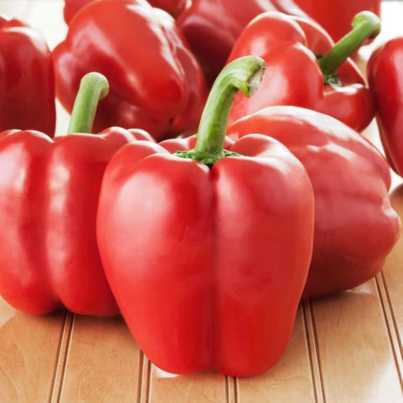 K&ouml;nnte beinhalten: Nahaufnahme mehrerer leuchtend roter Paprika mit gr&uuml;nen Stielen. Die Paprika sind auf einer hellbraunen Holzoberfl&auml;che angeordnet. Das Bild zeigt die glatte, gl&auml;nzende Haut und das frische Aussehen des Gem&uuml;ses.