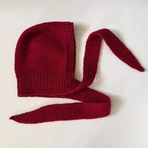 Può includere: Una cuffia lavorata a maglia di colore rosso intenso con una trama a coste lungo il bordo inferiore e due lunghi lacci affusolati che si estendono dai lati. Il copricapo è realizzato con un punto rasato uniforme su tutta la superficie.