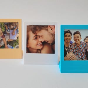 May include: Three different colored picture frames with different photos inside. The frames are all the same size and shape, but they are different colors: orange, white, and blue. The photos inside the frames are of different people and scenes.