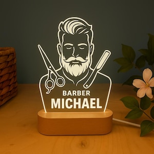 Puede incluir: Un letrero de barbería iluminado con una ilustración estilizada de barbero, tijeras y una navaja. El letrero está personalizado con el nombre "MICHAEL" y la palabra "BARBER". La base es de madera.