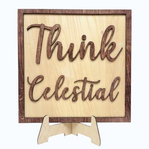 May include: Wooden sign with the words "Think Celestial" in a cursive font. The text is a dark brown color and is raised above the light-colored wood background. The sign has a dark brown frame and is supported by a wooden stand.