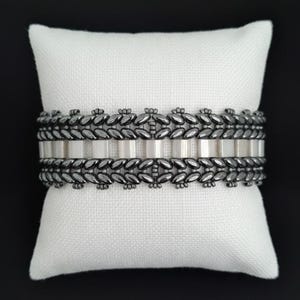May include: A silver and black beaded bracelet displayed on a white pillow against a black background. The bracelet features a repeating pattern of silver beads and black leaf-shaped accents, creating a textured design. The bracelet is approximately 0.75 inches wide.