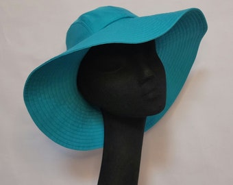 May include: A turquoise sun hat with a wide brim, displayed on a black mannequin head. The hat has a structured crown and a gently curved brim, offering ample shade. The hat's color is a vibrant, solid turquoise.