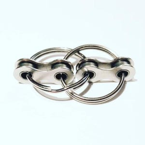 May include: Two silver-colored keychains, each featuring a bicycle chain link design. The keychains are interlocked, creating a unique and industrial aesthetic. The background is plain white.