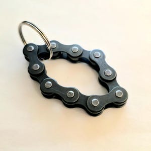 Peut inclure: Un porte-clés en forme de chaîne de vélo gris foncé avec un anneau argenté. La chaîne est disposée en forme de losange. Le porte-clés est en métal et a une finition mate. Le fond est de couleur beige clair.