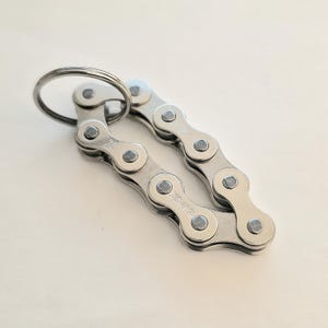 Puede incluir: Un llavero de cadena de bicicleta plateado con un anillo de metal. El llavero está hecho de eslabones de cadena conectados, con las letras "KMC" y "Z" visibles. Diseñado para llaves.