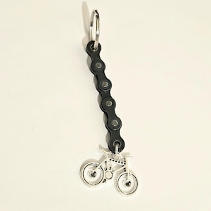 May include: A bicycle chain keyring with a silver-coloured bicycle charm. The keyring features a black bicycle chain with a silver key ring at the top. The bicycle charm is a detailed miniature bicycle.