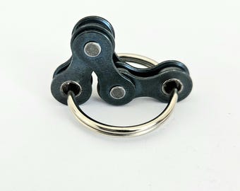 May include: A keychain featuring a bicycle chain design. The keychain is made of dark gray metal and includes two silver-colored key rings. The bicycle chain links are connected to form a unique and industrial-inspired accessory.