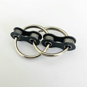 Peut inclure: Un puzzle en métal argenté avec deux anneaux imbriqués, chacun relié à une maillon de chaîne noir. Le puzzle est conçu pour être résolu en séparant les anneaux. Le fond est blanc.