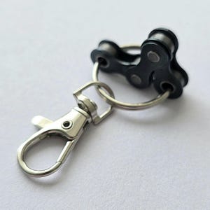 Peut inclure: Un porte-clés en chaîne de vélo noir avec un mousqueton et un porte-clés argentés. Le porte-clés présente un design unique, avec les maillons de la chaîne formant un élément décoratif. Le mousqueton est de style homard, et le porte-clés est rond.