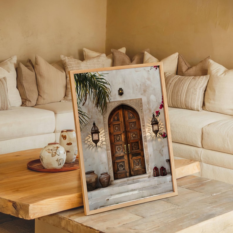 May include: A framed print of a photograph of a wooden door with intricate carvings. The door is set in a white wall with a palm tree in the background. Two lanterns hang on either side of the door.