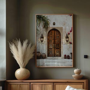 May include: A framed print of a photograph of a wooden door with intricate carvings. The door is set in a white wall with a palm tree in the background. There are two lanterns on either side of the door.