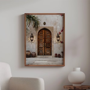 May include: A photograph of a wooden door with intricate carvings, framed by a white wall with a palm tree in the background. Two lanterns hang on either side of the door, and there are terracotta pots on the steps leading up to the door.