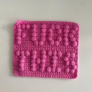 May include: A pink crocheted square dishcloth with a textured, raised pattern.