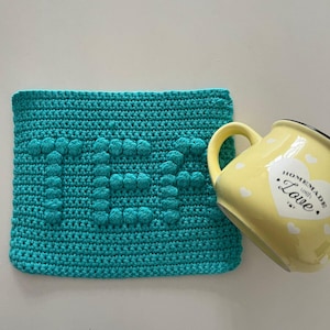 May include: A teal blue crocheted pot holder with the word "Love" in raised stitches. A yellow mug with white hearts and the text "Homemade with Love" is resting on the pot holder.
